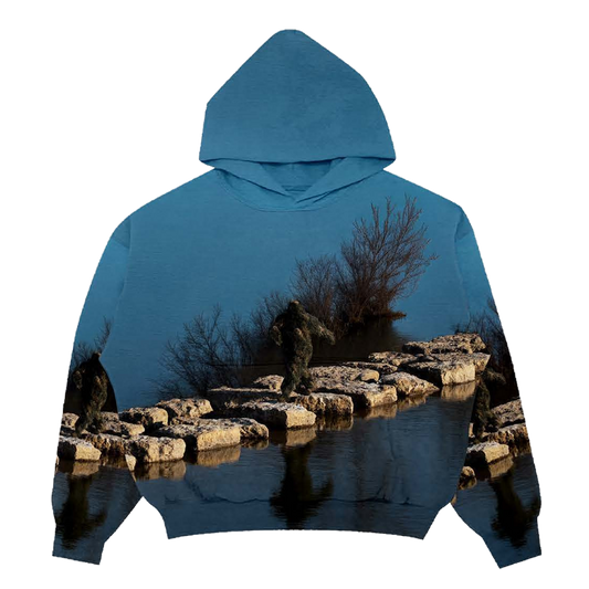 Hoodie with a nature-themed design of a Sasquatch waking on rocks in between a lake on a blue background