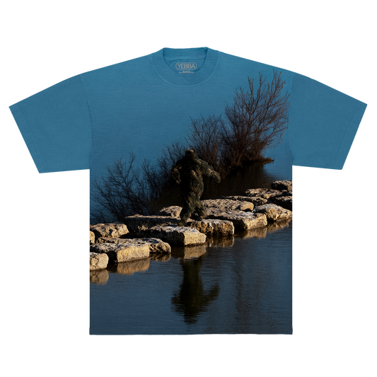 Blue shirt with a nature-themed design of a Sasquatch waking on rocks in between a lake on a blue background