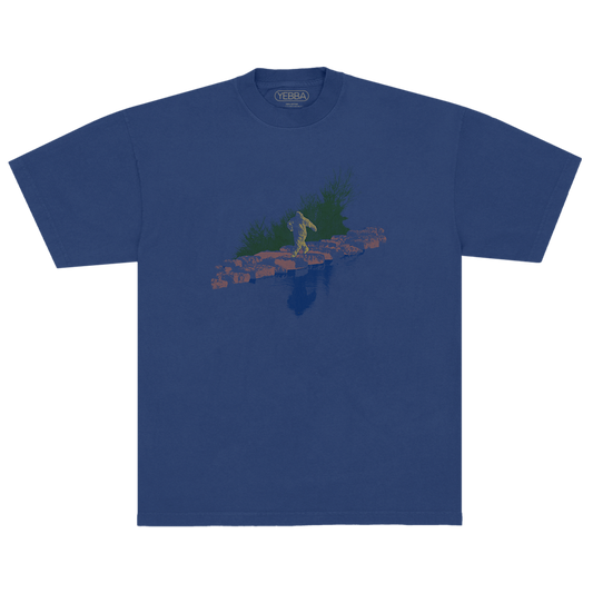 Blue t-shirt with a graphic design of a Sasquatch walking on rocks with grass behind it and a water refection in front. 